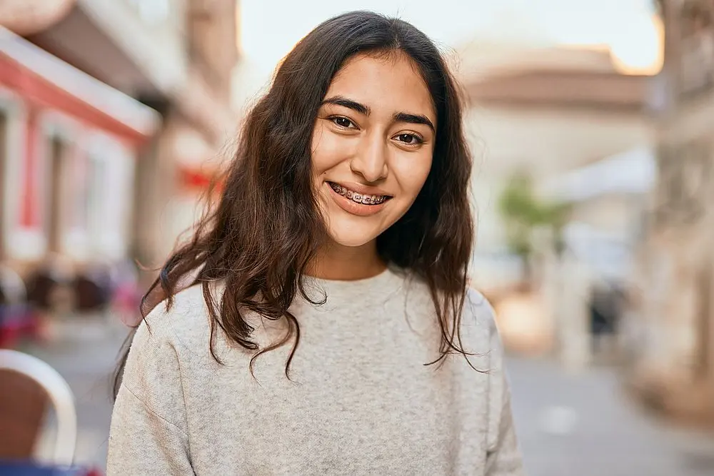Smiling outdoors in a light gray sweater, a young woman with long brown hair and braces shows off her confidence—one of the 5 proven benefits of Clear Color Braces for adults and children available at Koerich Orthodontics in Charlotte, Indian Trail or Monroe, NC: an attractive, natural look.