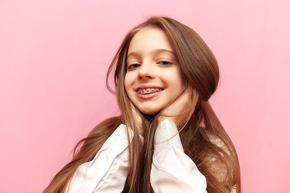 A young girl with long brown hair and braces, dressed in a white shirt, poses playfully against a pink background, possibly wondering, “Is chewing gum with braces bad for your teeth?” Koerich Orthodontics can help answer this and other orthodontic questions for patients in Charlotte, Indian Trail or Monroe, NC.
