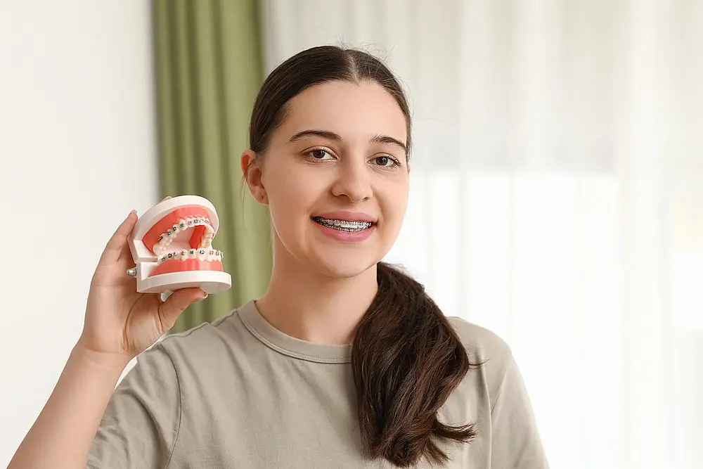 A teen girl with braces holds a dental model, illustrating concerns about crooked teeth—causes and straightening options. Koerich Orthodontics, in Charlotte, Indian Trail or Monroe, NC, can help address these issues. She is indoors with a green curtain and sheer white backdrop.