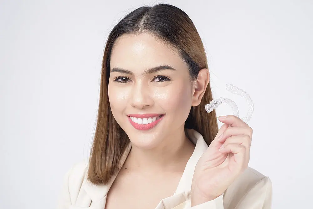 A woman with straight brown hair smiles and holds a clear dental aligner in her right hand, demonstrating how Invisalign works for Koerich Orthodontics in Charlotte, Indian Trail, or Monroe, NC.