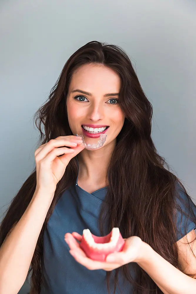 A woman with long brown hair confidently smiles while inserting a clear aligner into a dental model—showing how easy orthodontic care can be with Koerich Orthodontics in Charlotte, Indian Trail or Monroe, NC. If you ever lose an Invisalign tray, there’s no need to stress!.