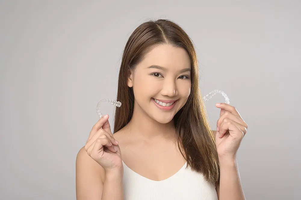 A woman with long brown hair smiles while holding clear orthodontic aligners against a plain background—an ideal image for Koerich Orthodontics in Charlotte, Indian Trail, or Monroe, NC to illustrate "How to Clean Invisalign Trays: The Complete Guide.