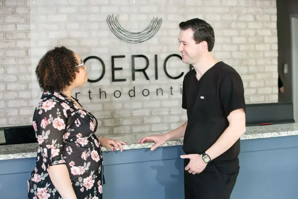 At the reception desk of Koerich Orthodontics in Charlotte, Indian Trail or Monroe NC, Dr. Leo talks with a patient while the Invisalign Indian Trail logo is visible on the wall behind them.