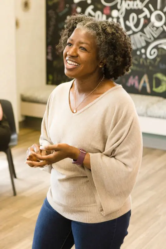 Inside Koerich Orthodontics in Charlotte, Indian Trail, or Monroe NC, a smiling woman with curly gray hair stands by a chalkboard wall, ready to share reviews and testimonials.