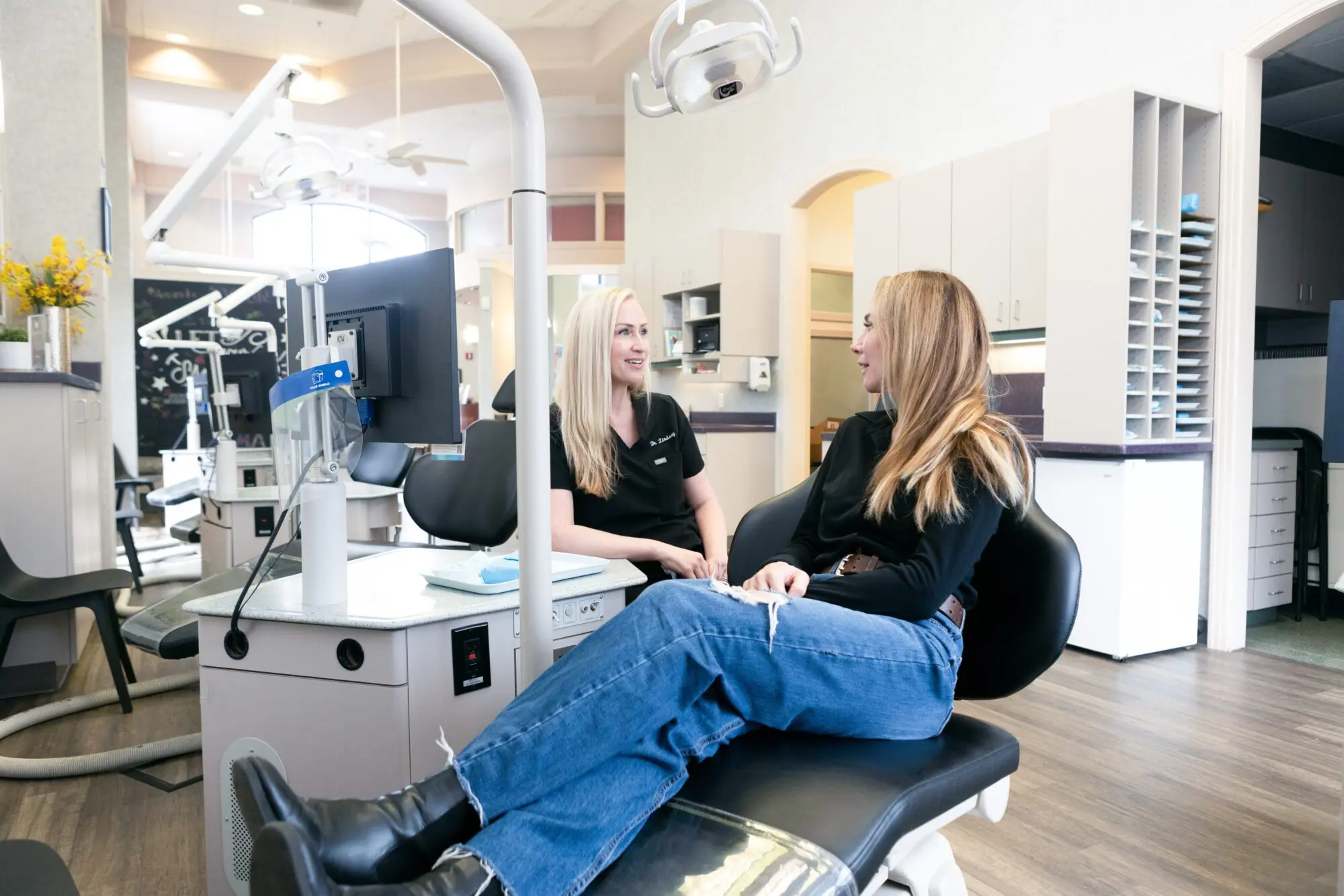 Two women are sitting in a dental office at Koerich Orthodontics. One is in a dental chair, chatting with the other beside her, surrounded by modern dental equipment in Indian Trail NC