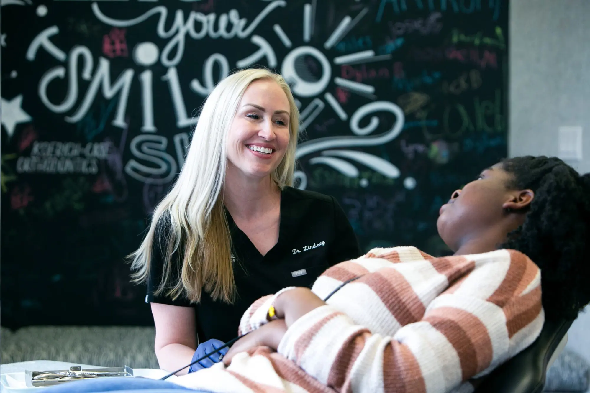 At Koerich Orthodontics in Charlotte, Indian Trail, or Monroe NC, a dentist in black scrubs smiles while speaking with a patient reclining in the dental chair, with a chalkboard featuring writing visible in the background.