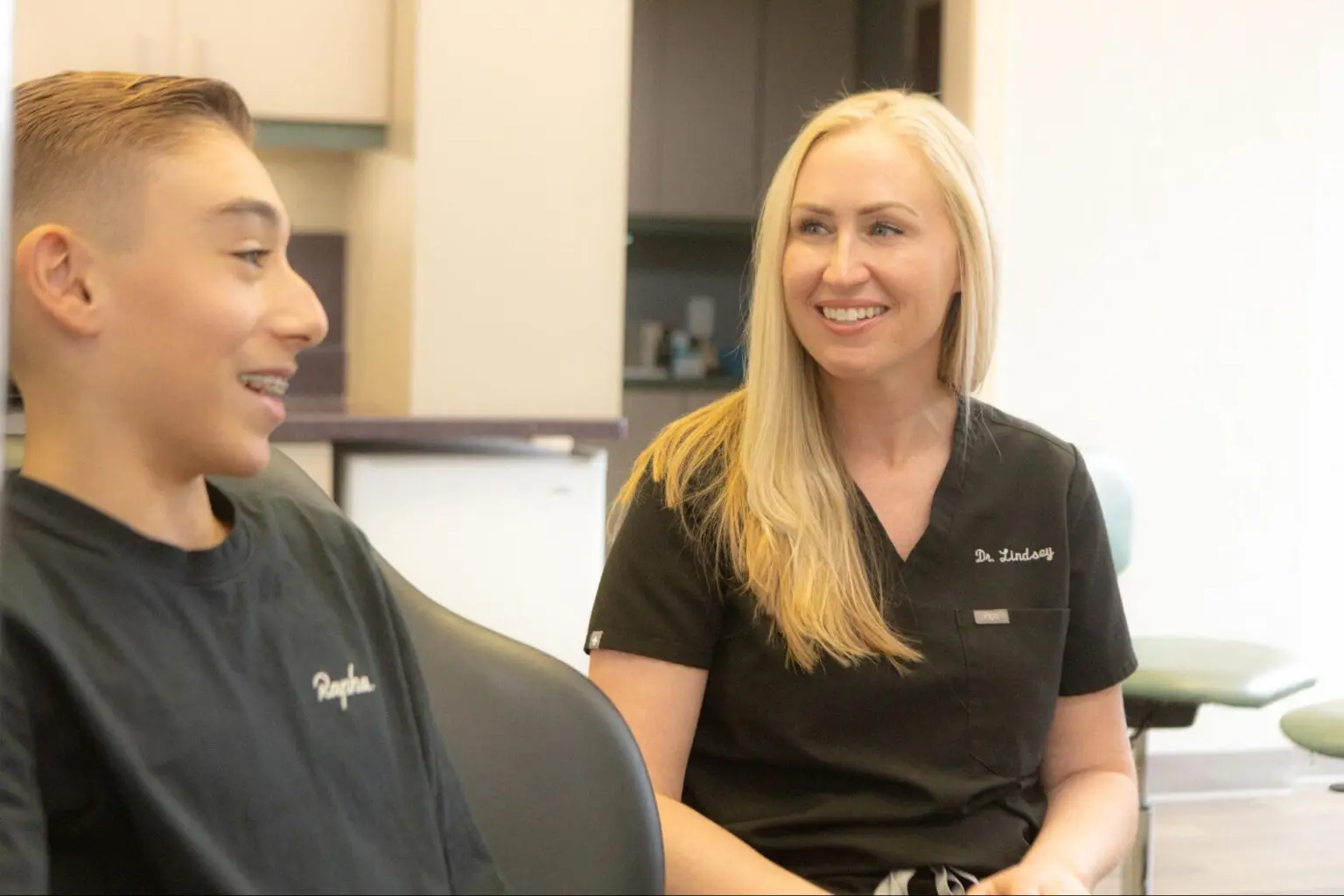 A young person with braces sits in a dental chair at Koerich Orthodontics, chatting with a smiling dentist who has long blonde hair and wears a sleek black uniform in Monroe NC