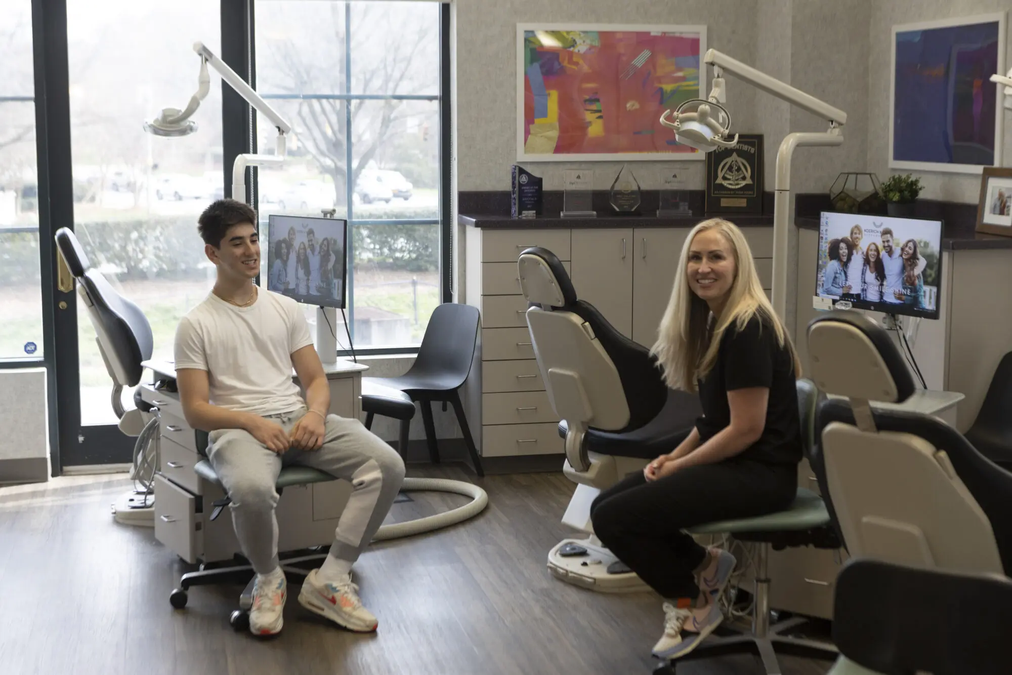 A young man and woman sit smiling at each other in dentist chairs at Koerich Orthodontics. Computer monitors in the background display group photos, adding a personal touch to the modern office ambiance in Indian Trail NC