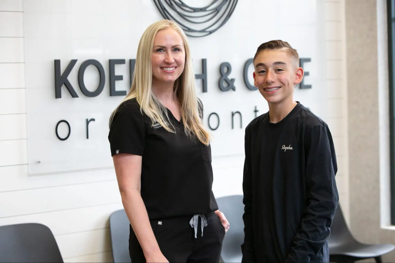 At Koerich Orthodontics in Charlotte, NC, a woman and a young person in black uniforms smile together in front of a sign that reads “Koehn & Sipe Orthodontics,” highlighting Dr. Lindsey Koerich’s expert orthodontic care.