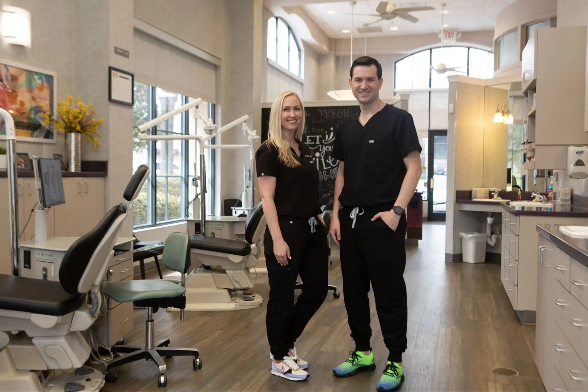 Two dental professionals in scrubs stand in a modern dental office at Koerich Orthodontics, smiling at the camera. Dental chairs and equipment are visible in the background, reflecting their commitment to exceptional care in Indian Trail NC