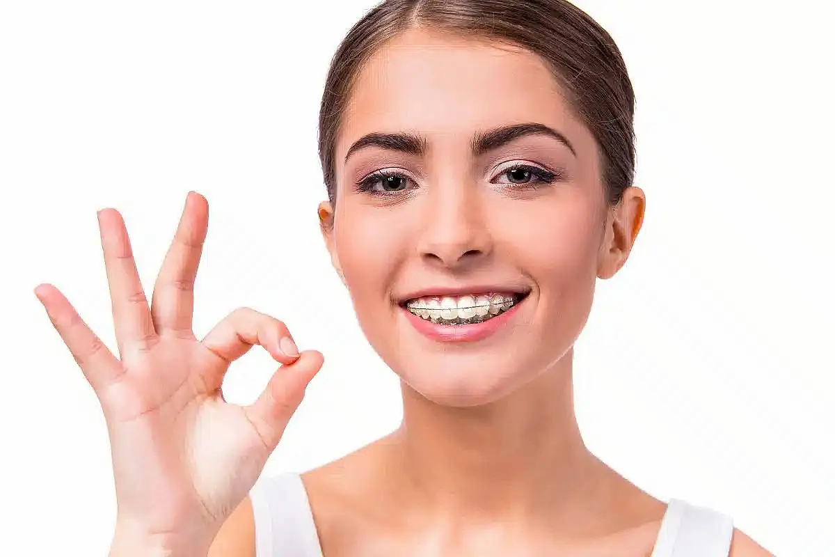 A woman with clear braces is smiling and making an "OK" gesture with her hand. She is wearing a white top against a neutral background in Charlotte, NC