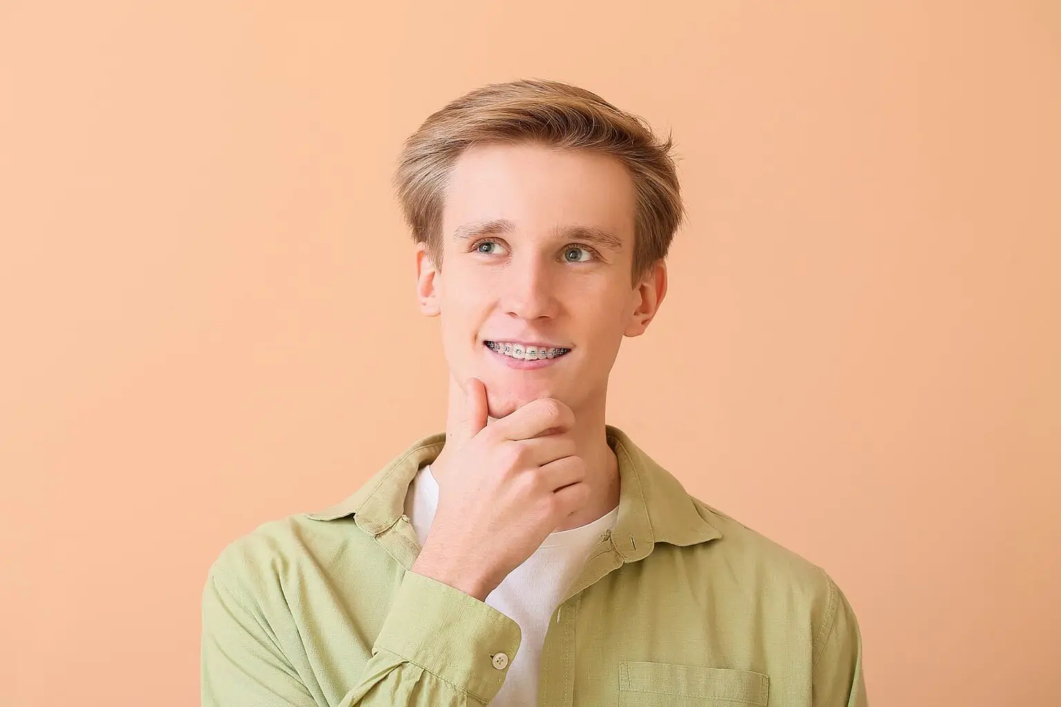 A young person with braces, dressed in a light green shirt and white undershirt, stands against a peach background, looking up thoughtfully and touching their chin—perhaps weighing aligners vs. braces at Koerich Orthodontics in Charlotte, NC.