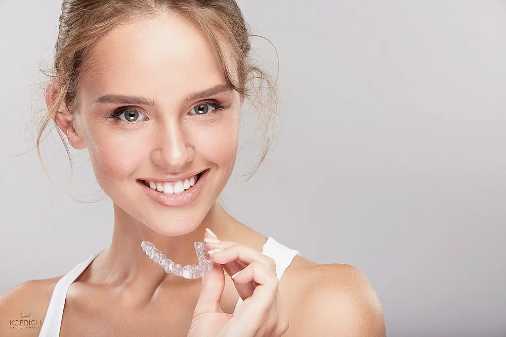 a blonde woman smiling while holding invisalign clear aligners after considering between aligners vs braces in Charlotte, NC