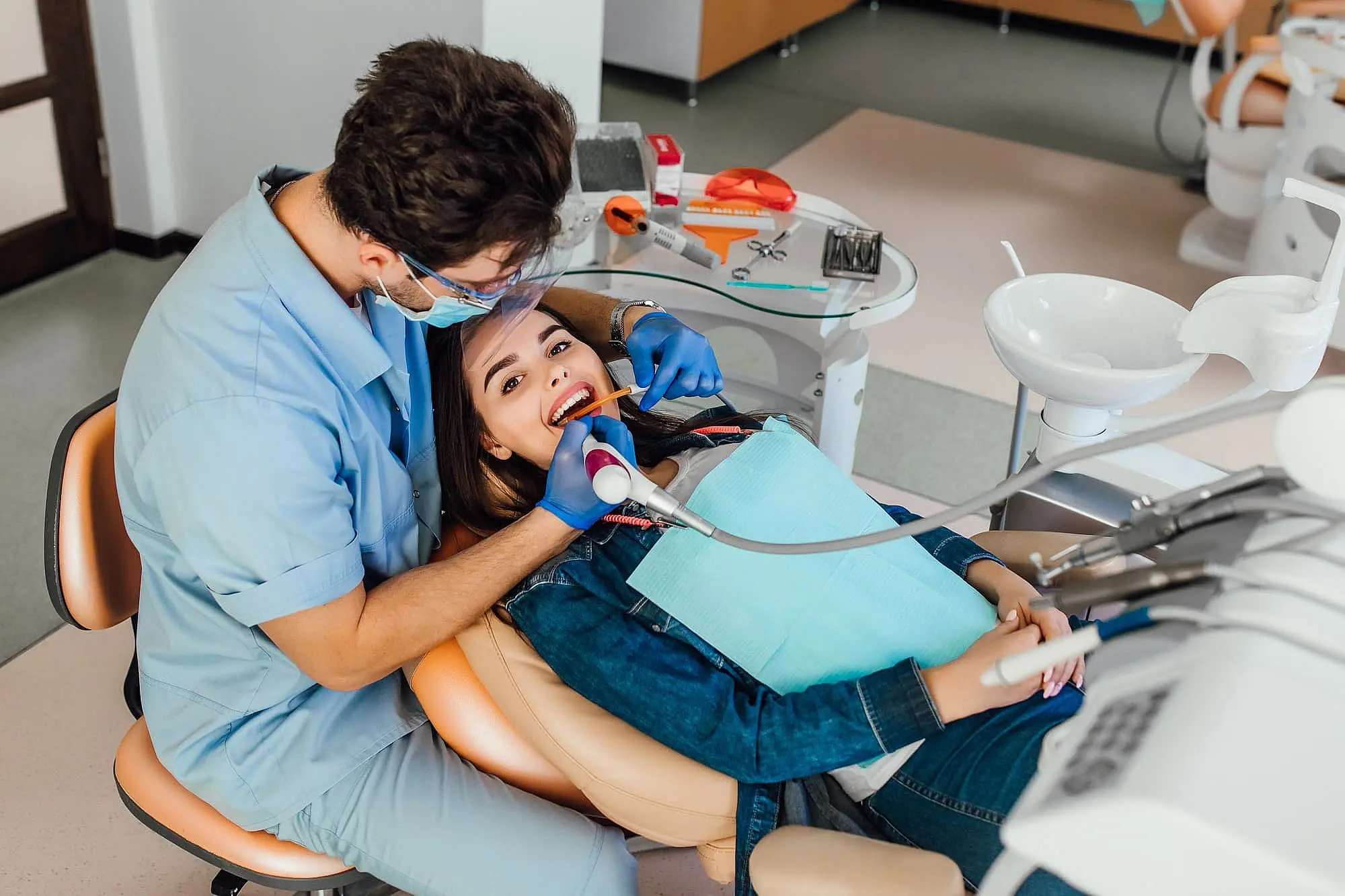 A dentist examines a smiling woman teeth in a dental office, where orthodontic appliances are neatly arranged nearby. The woman is wearing a green bib, while the dentist, holding dental tools, ensures everything is in place for optimal oral health in Charlotte, NC.