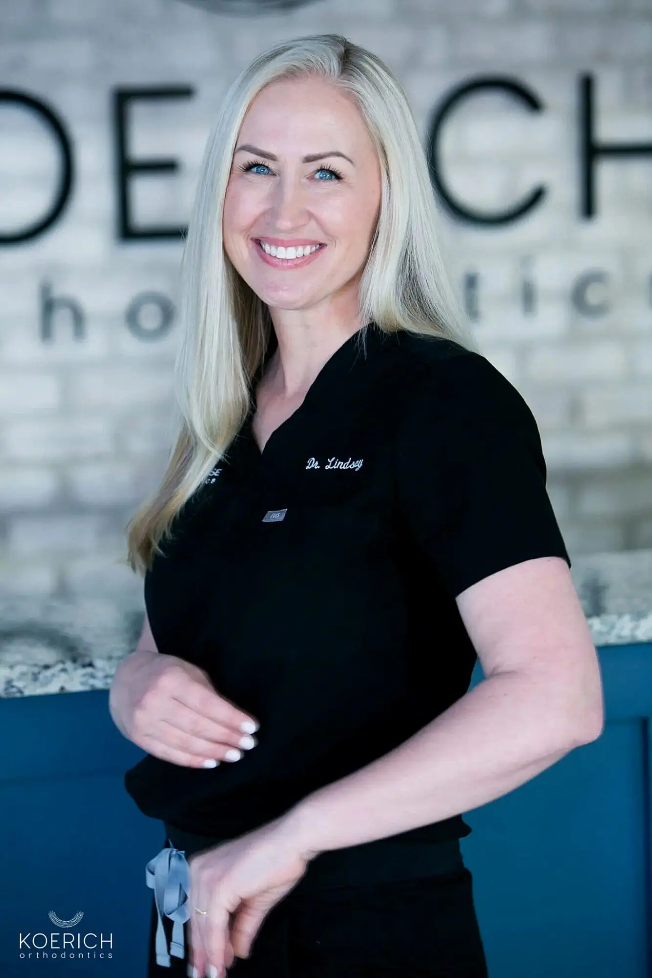 A person with long blonde hair smiles warmly, wearing a black professional uniform. In the background, a partially visible sign with large letters hints at an orthodontist practice in Charlotte in Charlotte NC