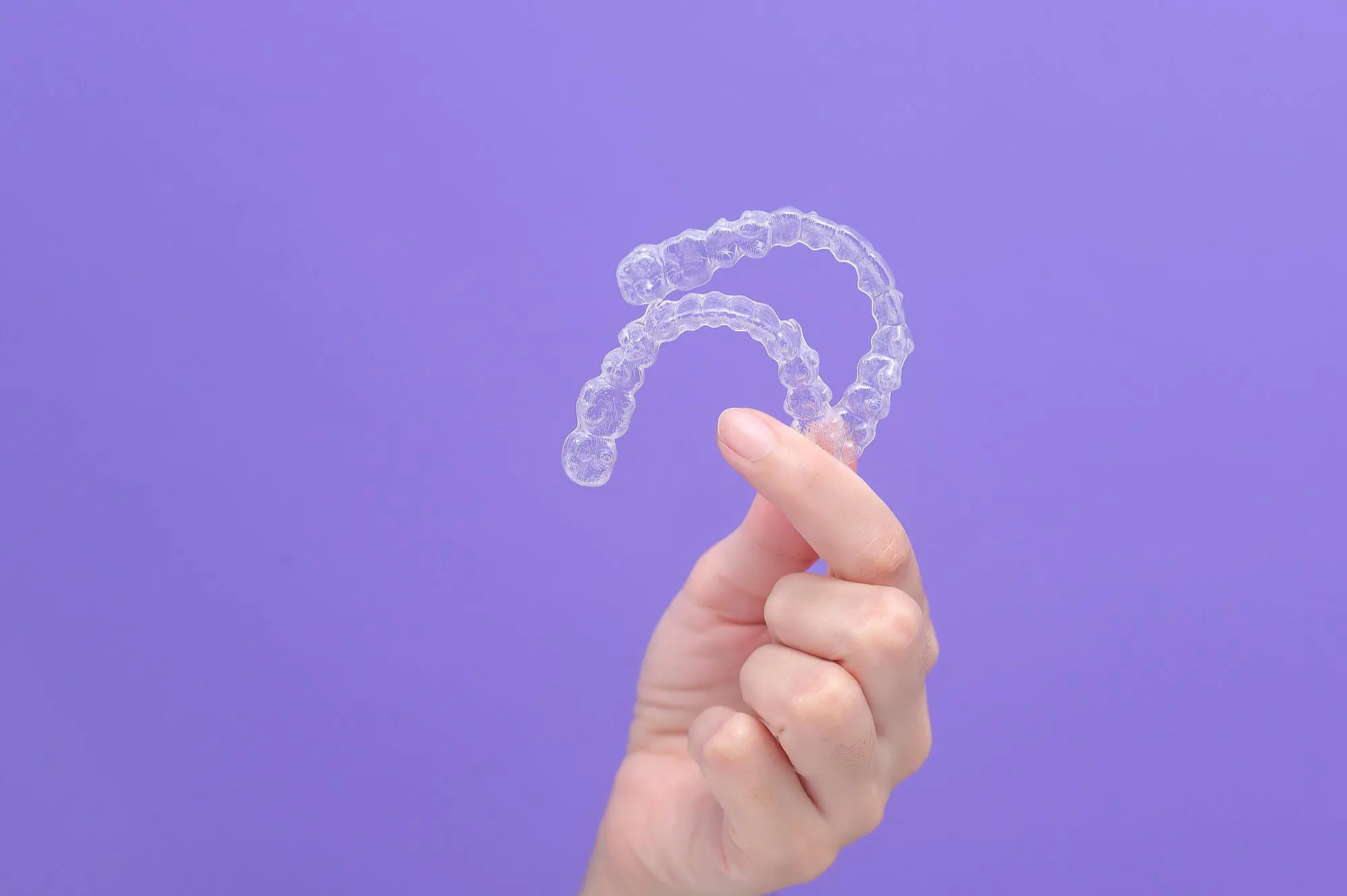 A hand displays two clear dental aligners against a solid purple background, showcasing the discreet design and value of Invisalign cost at Koerich Orthodontics in Charlotte, Indian Trail or Monroe, NC.