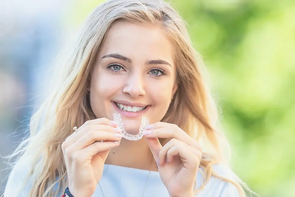 A woman with long blonde hair smiles outdoors, holding a clear dental aligner in both hands, as she thinks about does insurance cover Invisalign in Charlotte, NC at Koerich Orthodontics in Indian Trail or Monroe, NC.