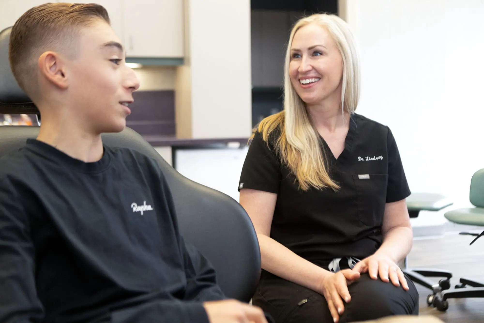 At Koerich Orthodontics in Ballantyne NC, a teen and a woman in black scrubs—likely an orthodontist—sit and talk in the office. The professional’s warm smile creates a welcoming healthcare environment.