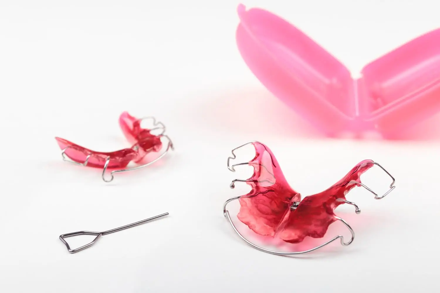 Displayed on a white surface are two pink orthodontic retainers, a metal adjustment tool, and an open pink plastic case—essential items for use with functional appliances recommended by Children’s Orthodontists at Koerich Orthodontics in Charlotte, Indian Trail and Monroe, NC.