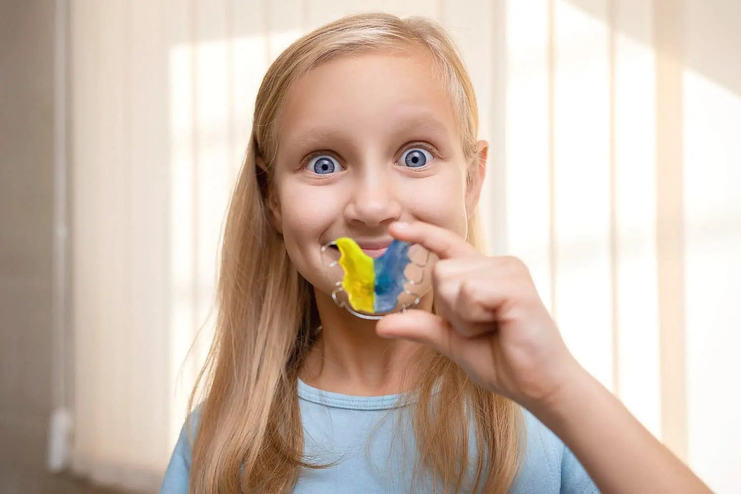 A young girl with long blonde hair proudly smiles indoors, showing off a colorful yellow-and-blue striped retainer—thanks to the expert care at Koerich Orthodontics in Charlotte, Indian Trail and Monroe, NC.
