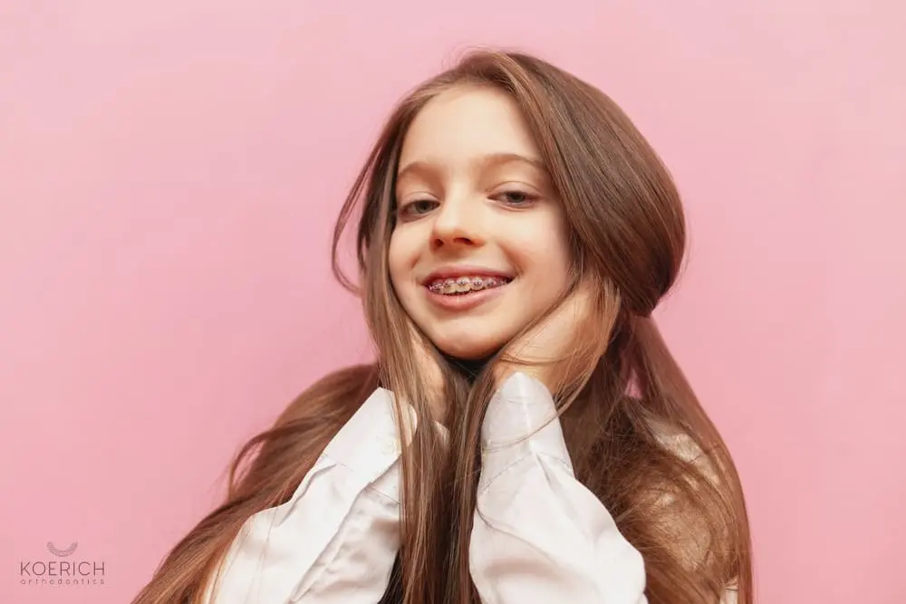A young girl with long brown hair and braces smiles with her hands on her cheeks against a pink background, representing Koerich Orthodontics' braces for overbite in Charlotte, Indian Trail or Monroe, NC.