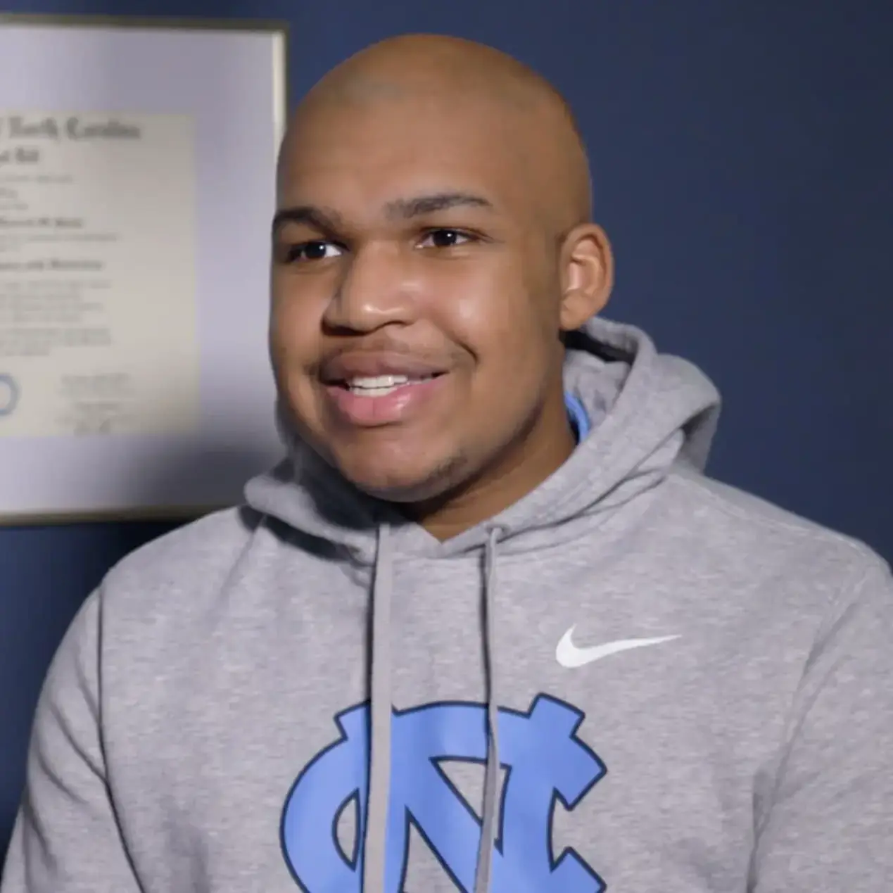 At Koerich Orthodontics in Charlotte, Indian Trail, or Monroe NC, a person in a gray hoodie with a blue "NC" logo smiles indoors, with a framed certificate and orthodontic patient testimonials visible on the wall behind them.