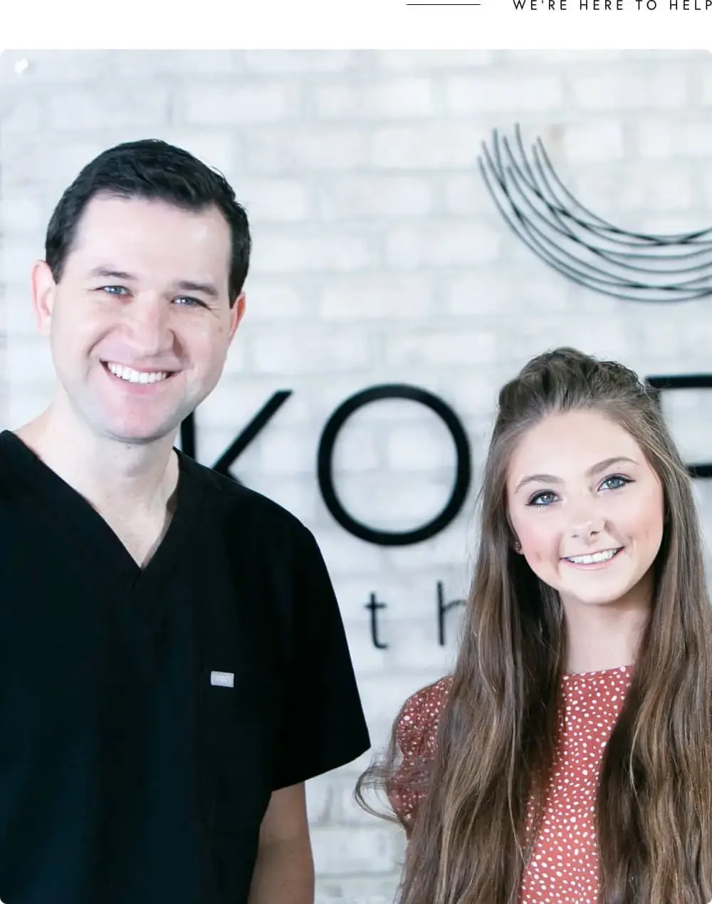 At Koerich Orthodontics in Charlotte, Indian Trail, or Monroe NC, a man in black scrubs and a woman in a red blouse with long hair stand smiling before their logo wall, showcasing their dedication to Same-Day Treatment.