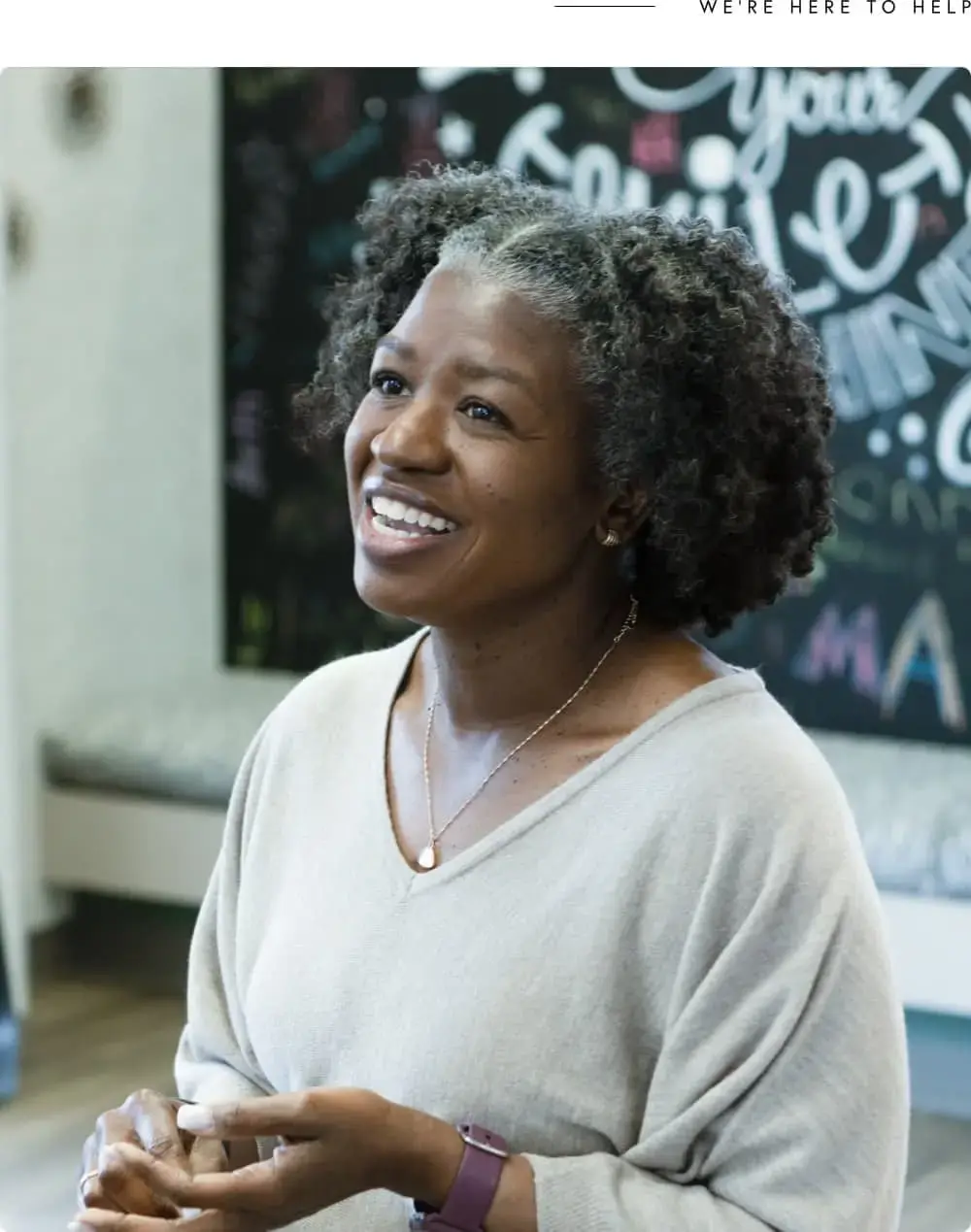 At Koerich Orthodontics in Charlotte, Indian Trail or Monroe NC, a woman with gray curly hair, dressed in a light sweater and necklace, smiles indoors before a blackboard displaying colorful payment plan information.