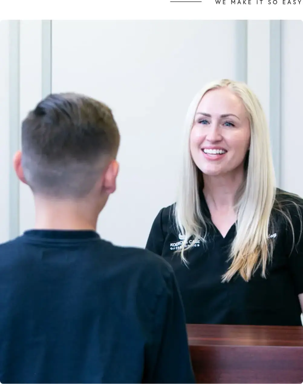 At Koerich Orthodontics in Charlotte, Indian Trail, or Monroe NC, a woman in a black shirt warmly smiles and talks with a boy across the counter as they get ready to begin treatment.