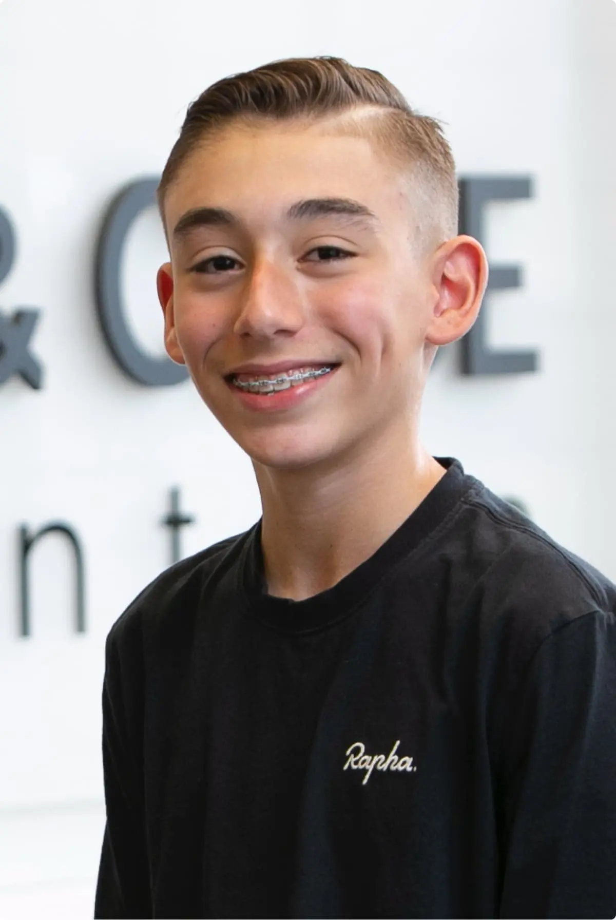 A teenage boy with short brown hair and braces, dressed in a black "Rapha" T-shirt, smiles at the camera against a blurred indoor background. At Koerich Orthodontics in Charlotte, Indian Trail or Monroe NC, it all starts with a smile!.