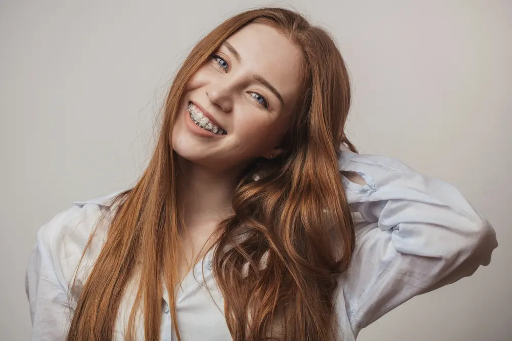 Smiling young woman with red hair and braces holds her hair back, representing Koerich Orthodontics in Charlotte, Indian Trail or Monroe, NC - Clear Braces vs Metal Braces in Charlotte, NC