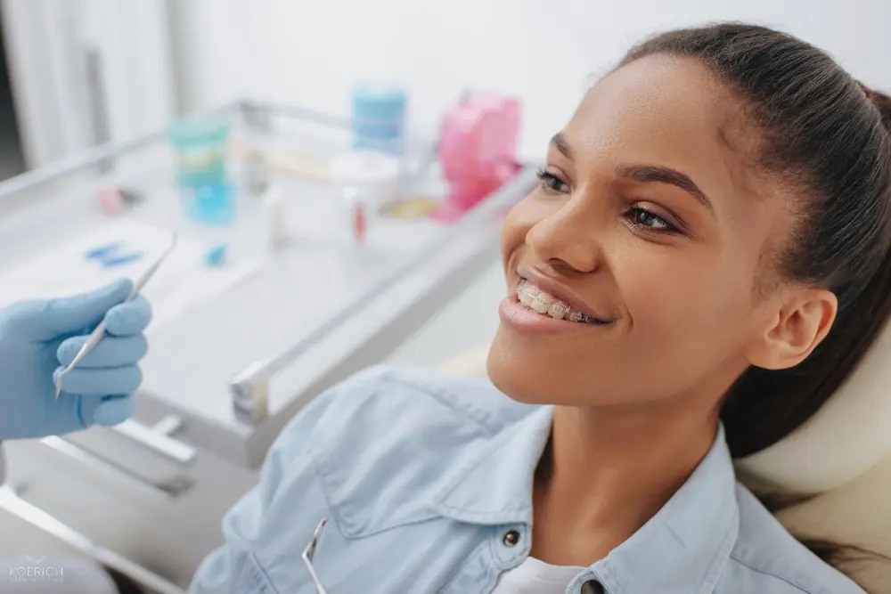 At Koerich Orthodontics in Charlotte, Indian Trail or Monroe, NC, a young woman with braces smiles in a dental chair by a dentist - Clear Braces vs Metal Braces in Charlotte, NC
