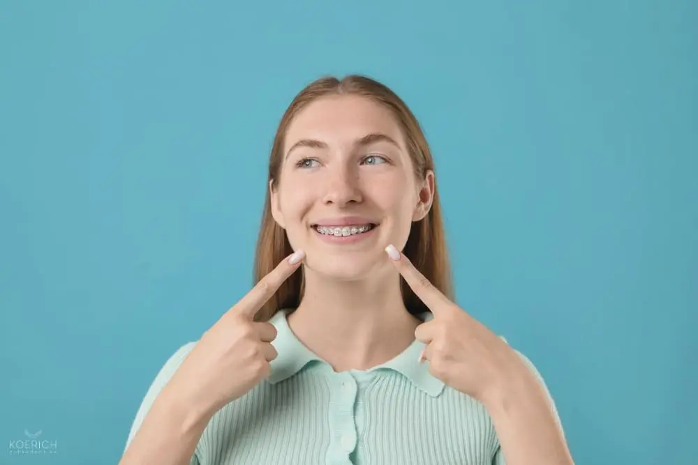 Smiling young woman with braces points at her teeth; Koerich Orthodontics in Charlotte, Indian Trail or Monroe, NC. Blue background - How to Clean Braces in Charlotte, NC 