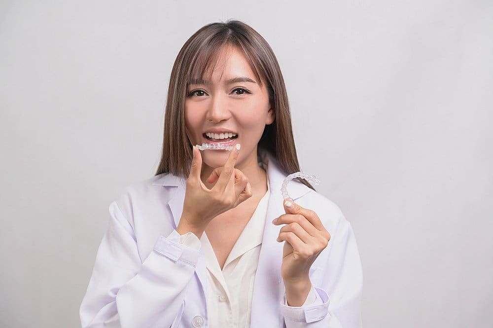 A woman in a white coat holds and points to a clear dental aligner, highlighting one of the popular alternatives to braces in Charlotte and Indian Trails, NC, against a plain background.